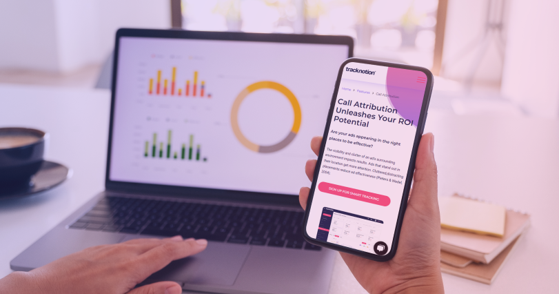 a Mobile phone showing a TrackNotion article on attribution, held in front of a laptop screen with analytics data.